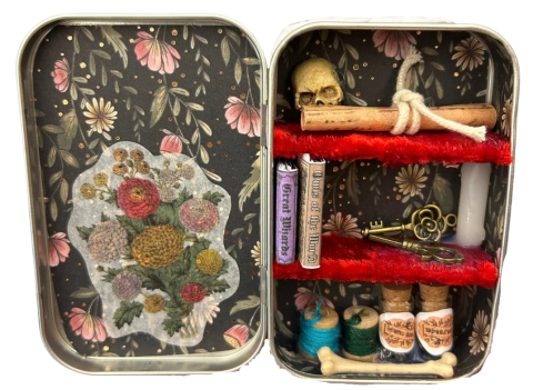 Open Altoid-size tin, inside left: paper with floral print on black background and a bunch of flowers on a white background over it at the bottom; inside right: the same black floral print inside right two shelves covered in fuzzy red material with mini skull and scroll on top shelf; two mini books, a mini brass key, and a mini white candle on middle shelf; and two mini spools of thread, two mini jars with labels, and a bone taking up the width in front on bottom 